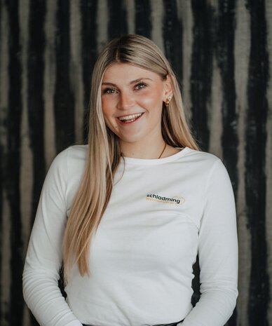 Lächelnde Frau mit langen blonden Haaren, weißes Shirt mit Logo schladming