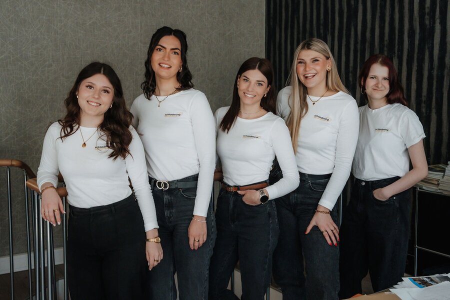 Five young women smiling in white shirts and dark jeans indoors