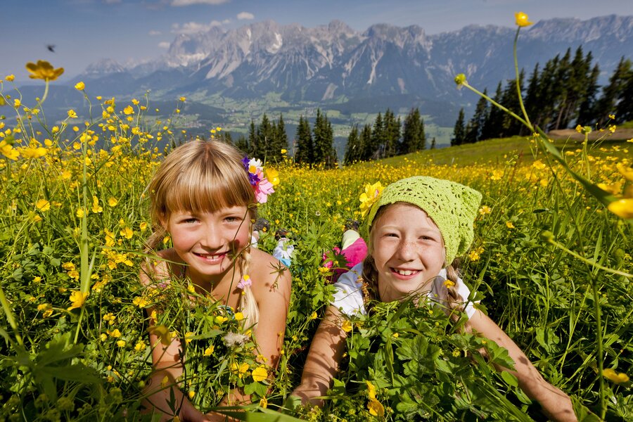 Two smiling girls lie in a yellow flower field with mountains.