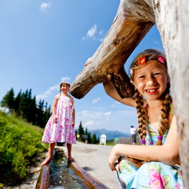 Two girls outdoors, one smiling with braided hair near a large log. | © Tom Lamm
