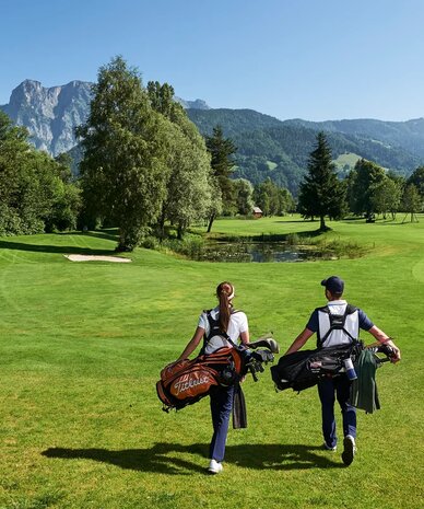 Two golfers walk across a sunny green golf course toward distant mountains