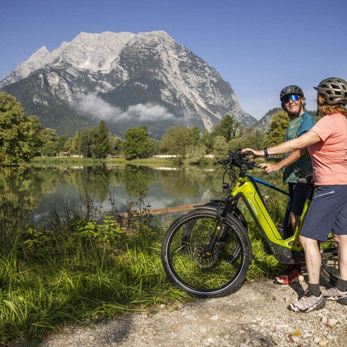 Two helmeted cyclists beside a lime electric bike by a lake.