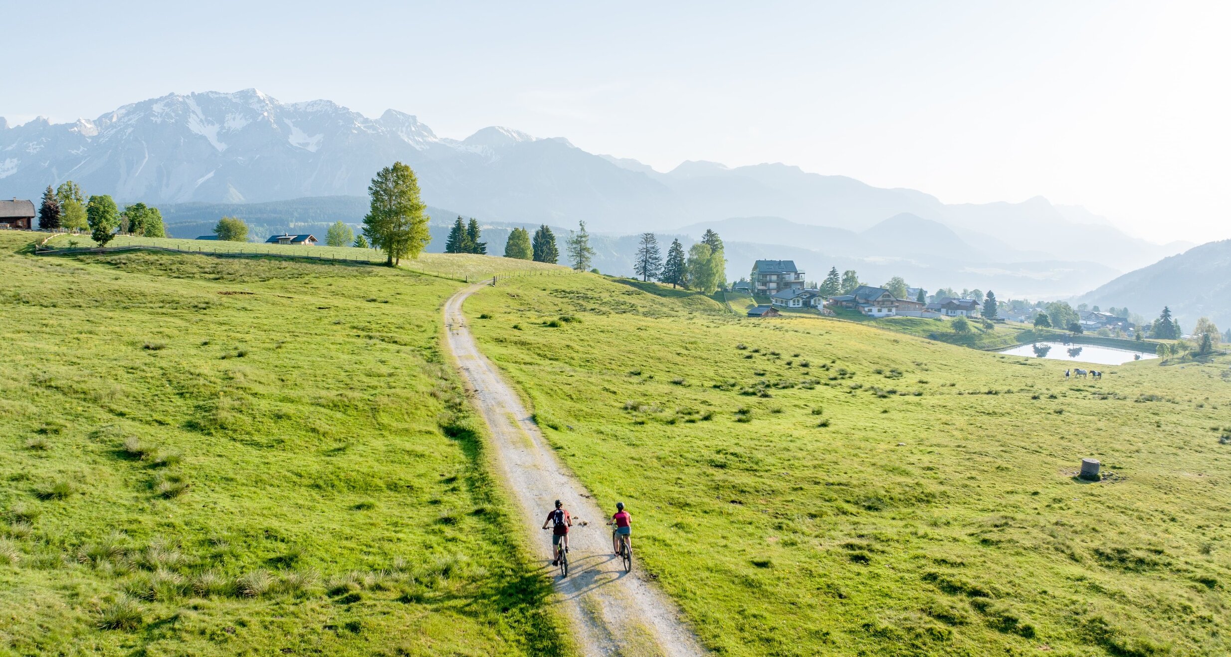 Zwei Radfahrer fahren auf einem Schotterweg durch grüne Weiden mit entfernten Bergen