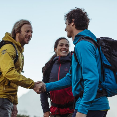 Drei Wanderer mit Rucksäcken begrüßen sich in bergiger Landschaft | © Sebastian Stiphout
