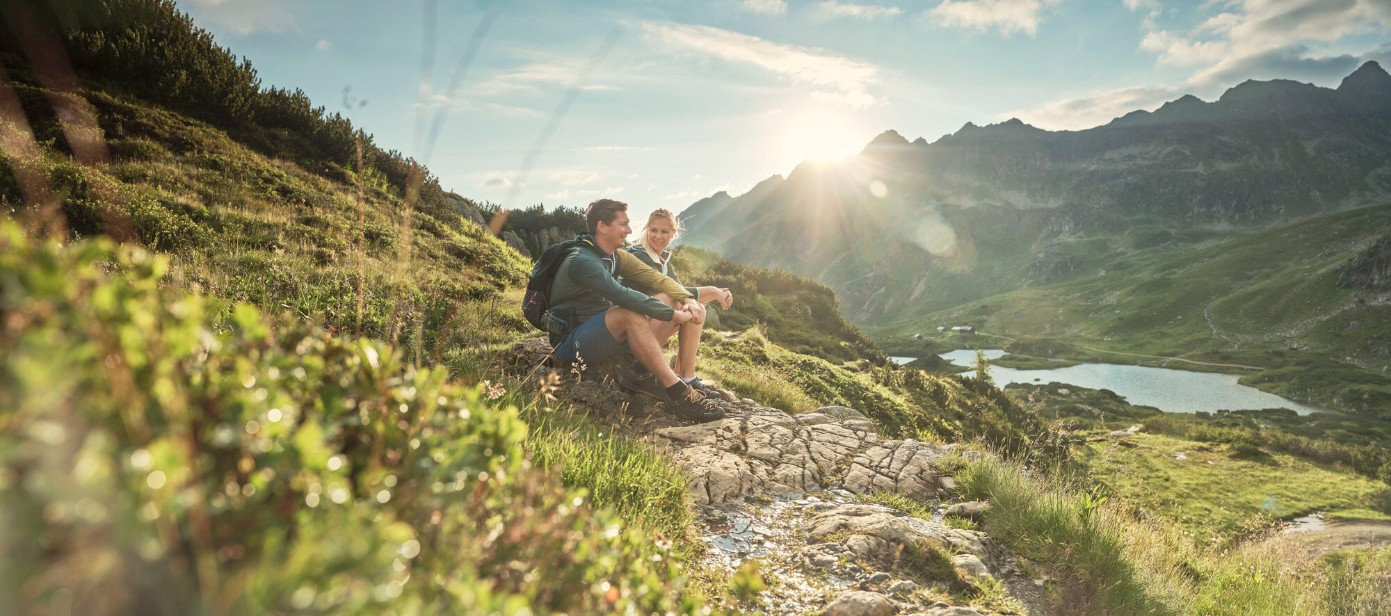Two hikers rest on a rocky slope overlooking alpine valley