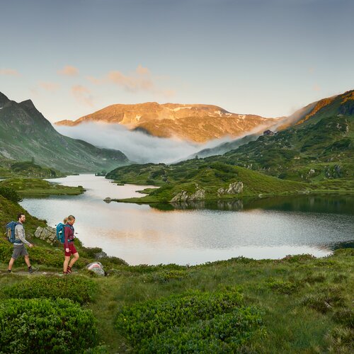 Two hikers with backpacks walk along alpine trail beside a lake.
