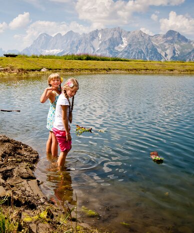 Two girls stand in a shallow alpine lake by a grassy bank.