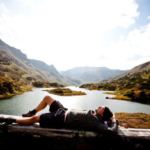 A person lying on a wooden bench by a mountain lake