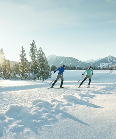 Two skiers glide across a snowy landscape toward forested mountains. | © Peter Burgstaller