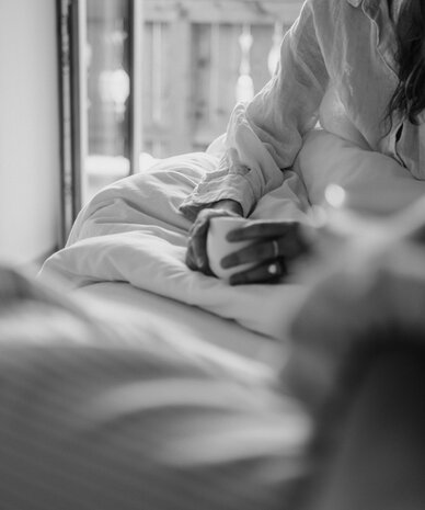 Person sitzt aufrecht im Bett und hält eine Tasse am Fenster.