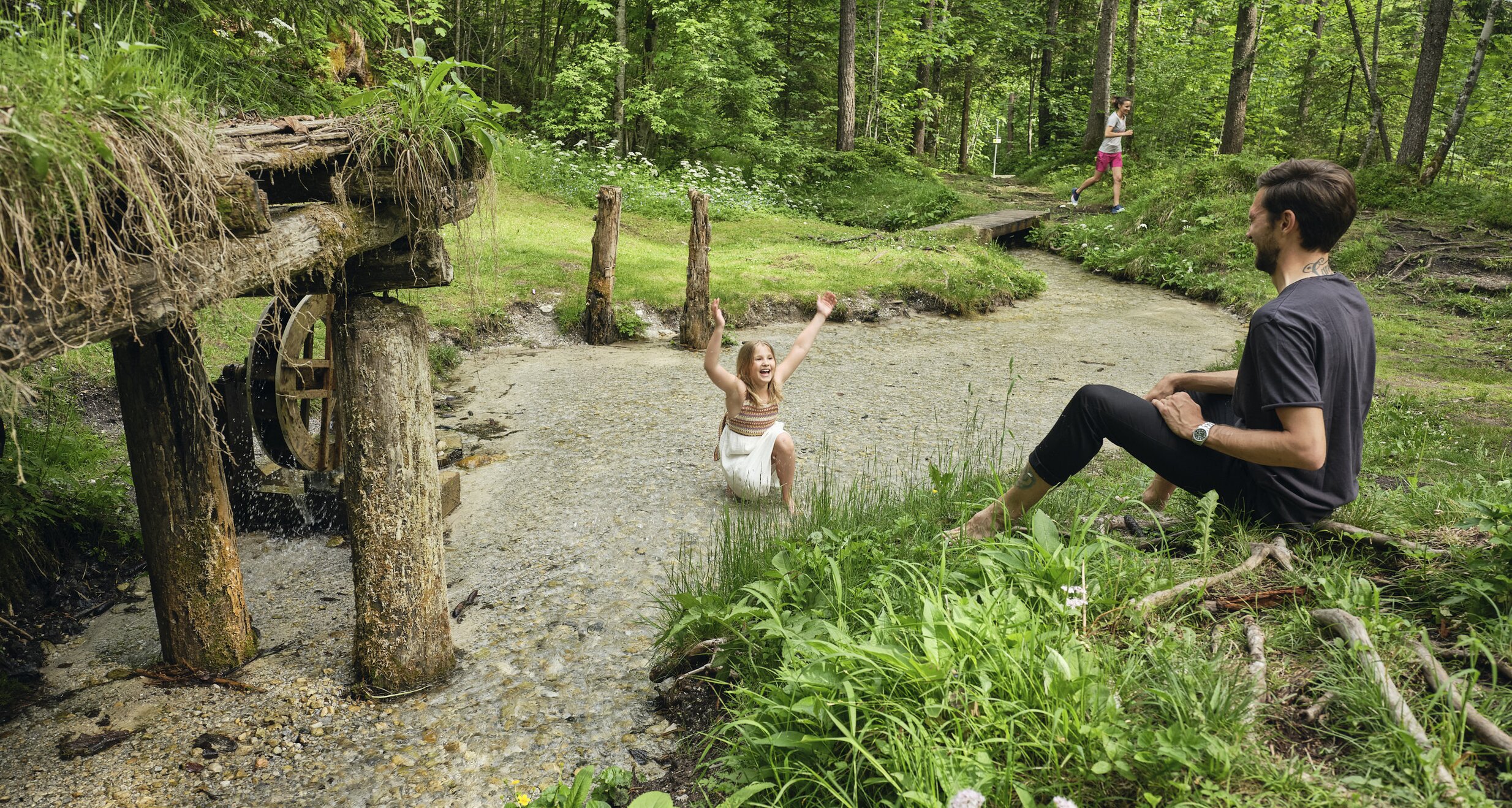 Girl splashes in shallow stream while man sits nearby. | © RAPHAELGABAUER.COM