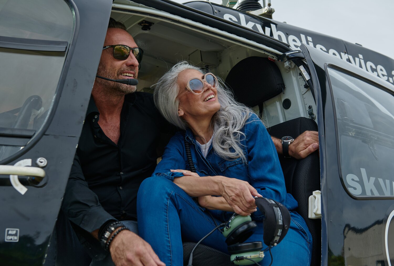 Two adults wearing headsets sit inside a helicopter cockpit, smiling.