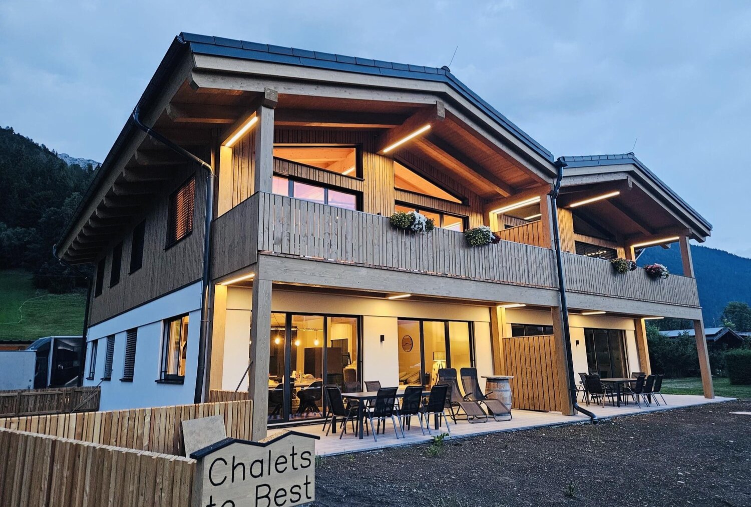 Zweistöckiges Holz-Chalet mit Balkon und Außenbestuhlung bei Dämmerung.
