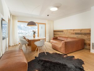 Wohnzimmer mit Ledersofa, Holzwand, Esszimmertisch und großem Fenster.