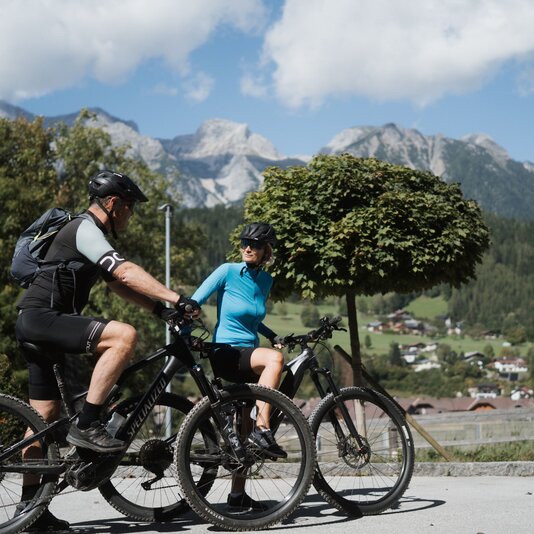 Two helmeted cyclists ride mountain bikes on a scenic road.